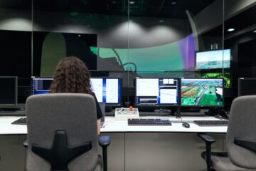 Mulher de cabelo cacheado, de costas em frente a vários computadores